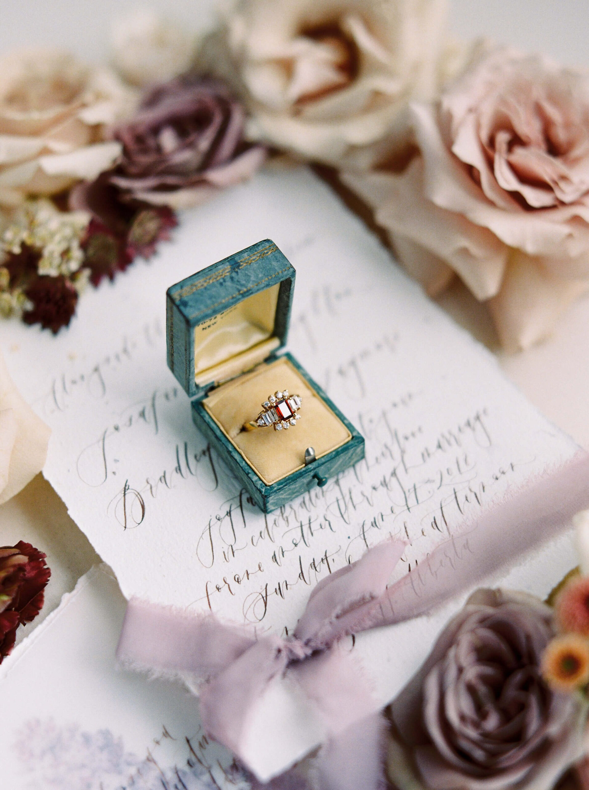 Why Wedding Details are So Important - A teal ring box sitting on top of a hand written invitation wrapped with silk ribbon and surrounded by soft pink roses