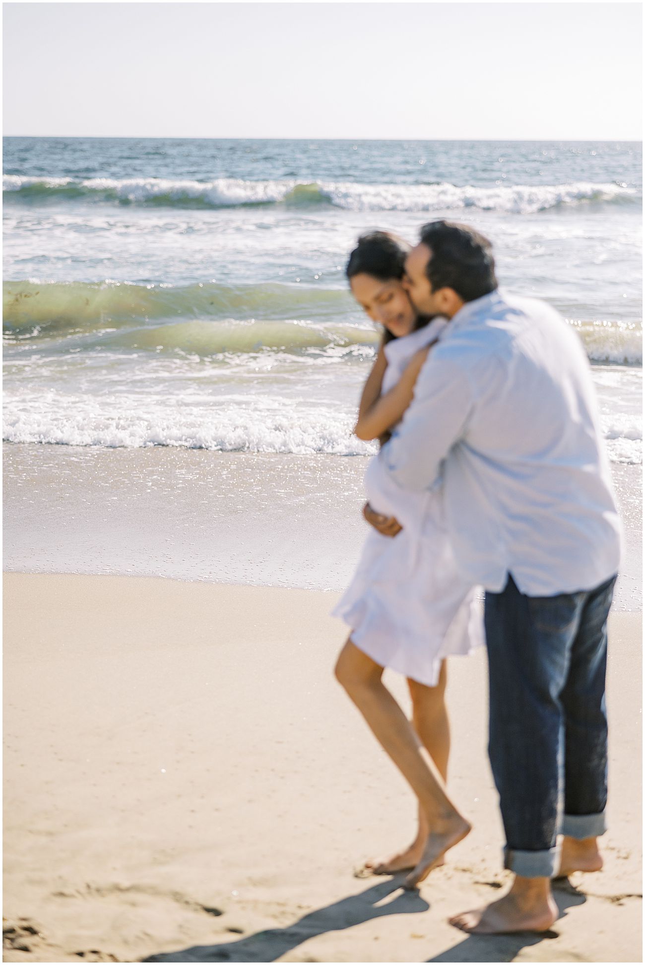 Husband hugging his pregnant wife from behind with the ocean in the background. She's wearing a white dress and he's wearing a blue button down and jeans Manhattan Beach maternity photographer