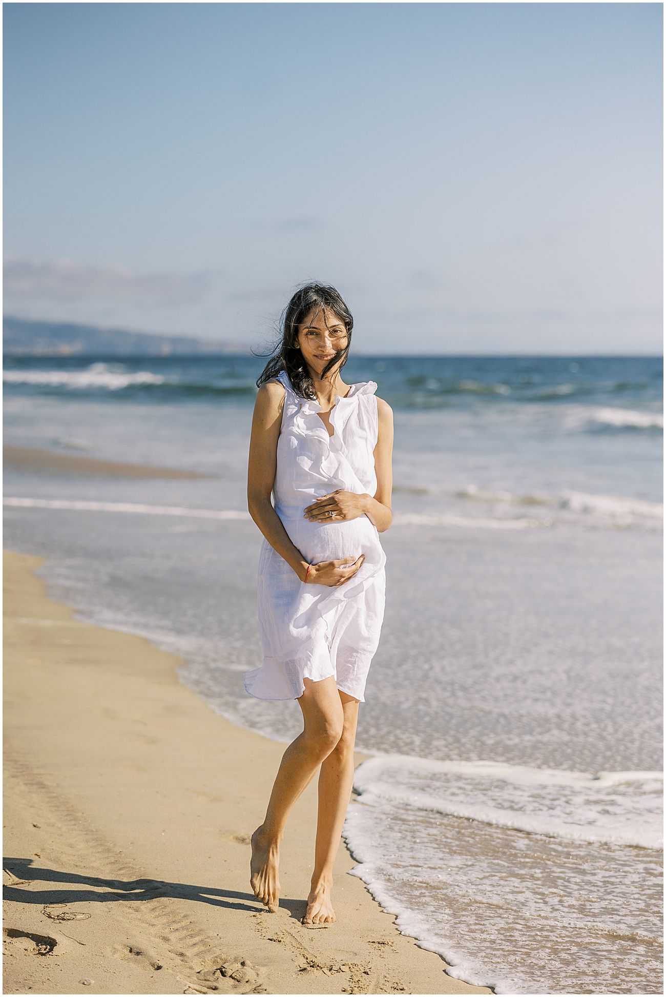 pregnant woman wearing a white dress being photographed at manhattan beach in southern california