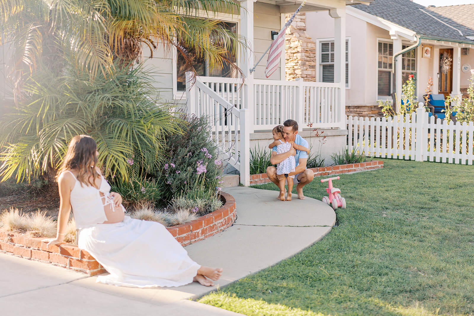 Pregnant woman sitting on a brick wall watching her husband hug their toddler in their front yard 5 Reasons Choose In-Home Maternity Photos in Orange County, California